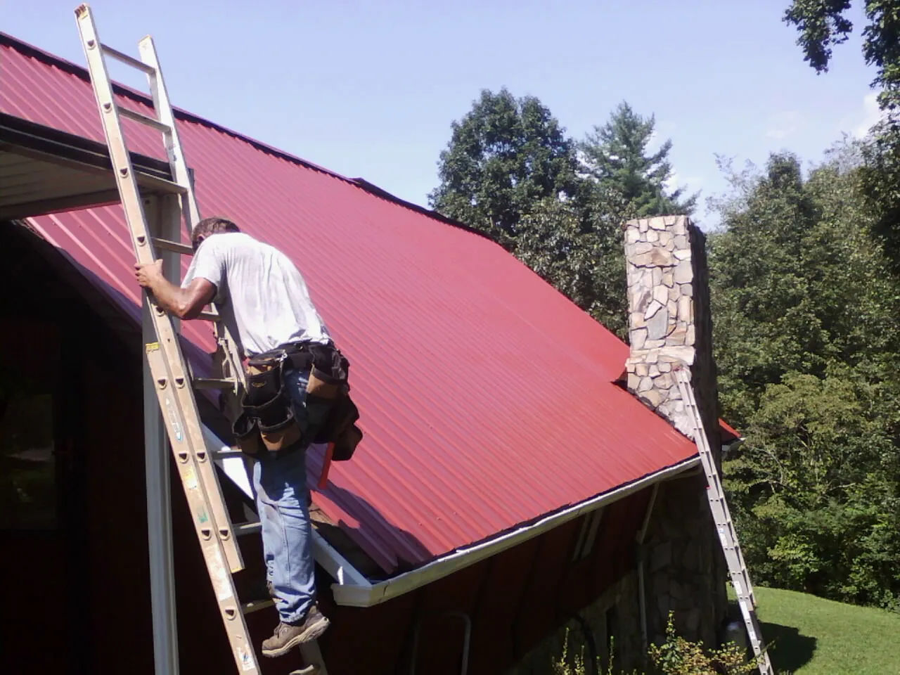 Roofer on a ladder beside a red metal roof and stone chimney — Soffit & Fascia Repair by Roofing Brothers in Braceville